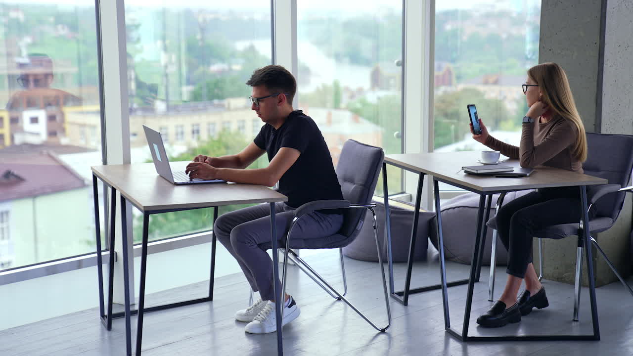 Young people working in the light spacious office. Man and woman sitting at desks, male typing on laptop and female colleague using smartphone. City view in window at backdrop in blur.