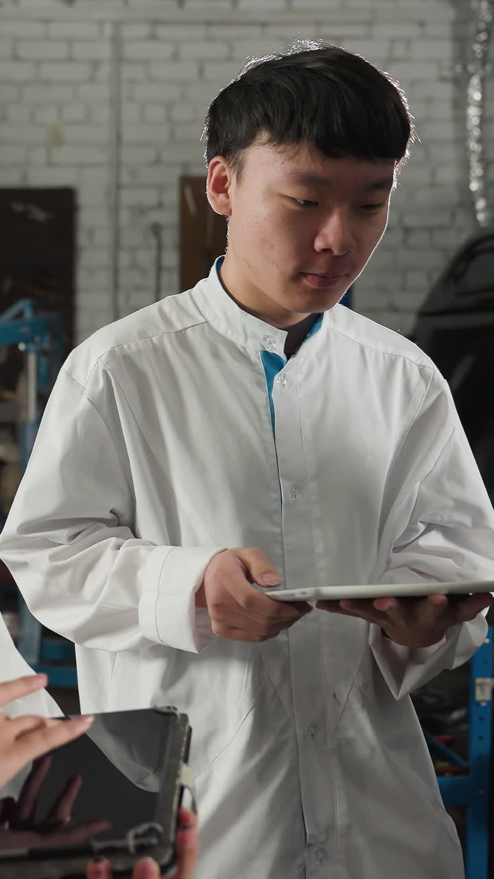 기계공이 자동차 작업장에서 학생들에게 볼트 풀기를 보여주고, 색 실험실 코트를 입은 두 명의 학생이 태블릿에 메모를 취하는 동안 다른 동료가 관찰하고, 배경에는 도구가 있습니다.