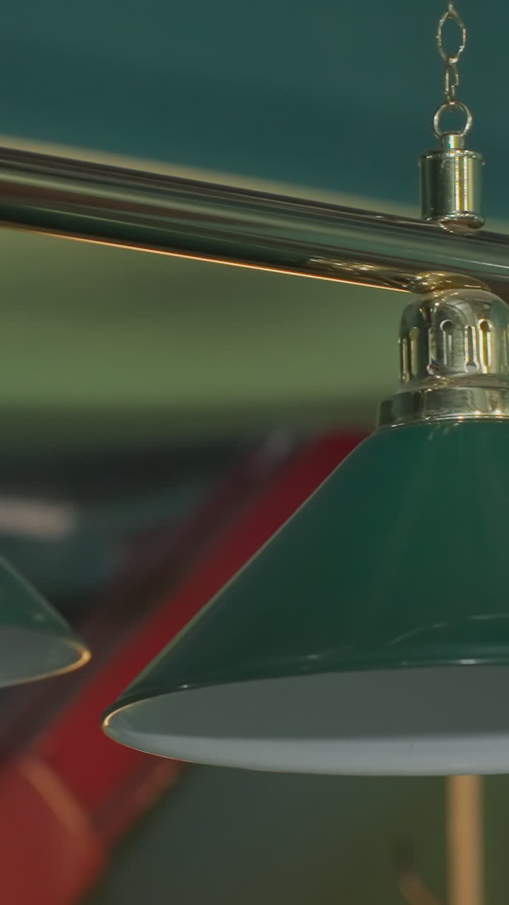 Nærbillede af to lamper over hovedet med grønne skærme i et billardrum, der fremhæver deres gyldne metalliske detaljer og elegante design, og kaster blødt lys på poolbordet nedenunder