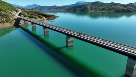 areal shot of motorcycles crossing a long empty bridge over a calm blue lake near the hills
