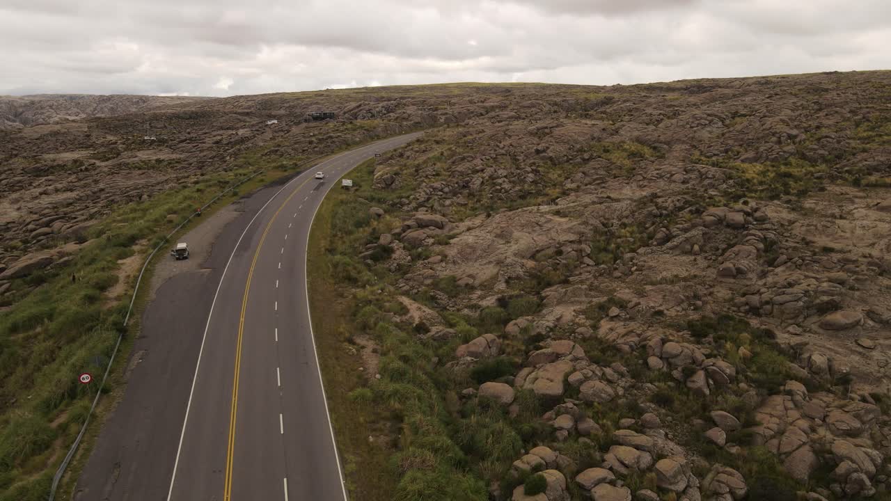 siguiente toma aérea de un automóvil blanco conduciendo por un camino rural rodeado de un paisaje montañoso argentino durante un día nublado