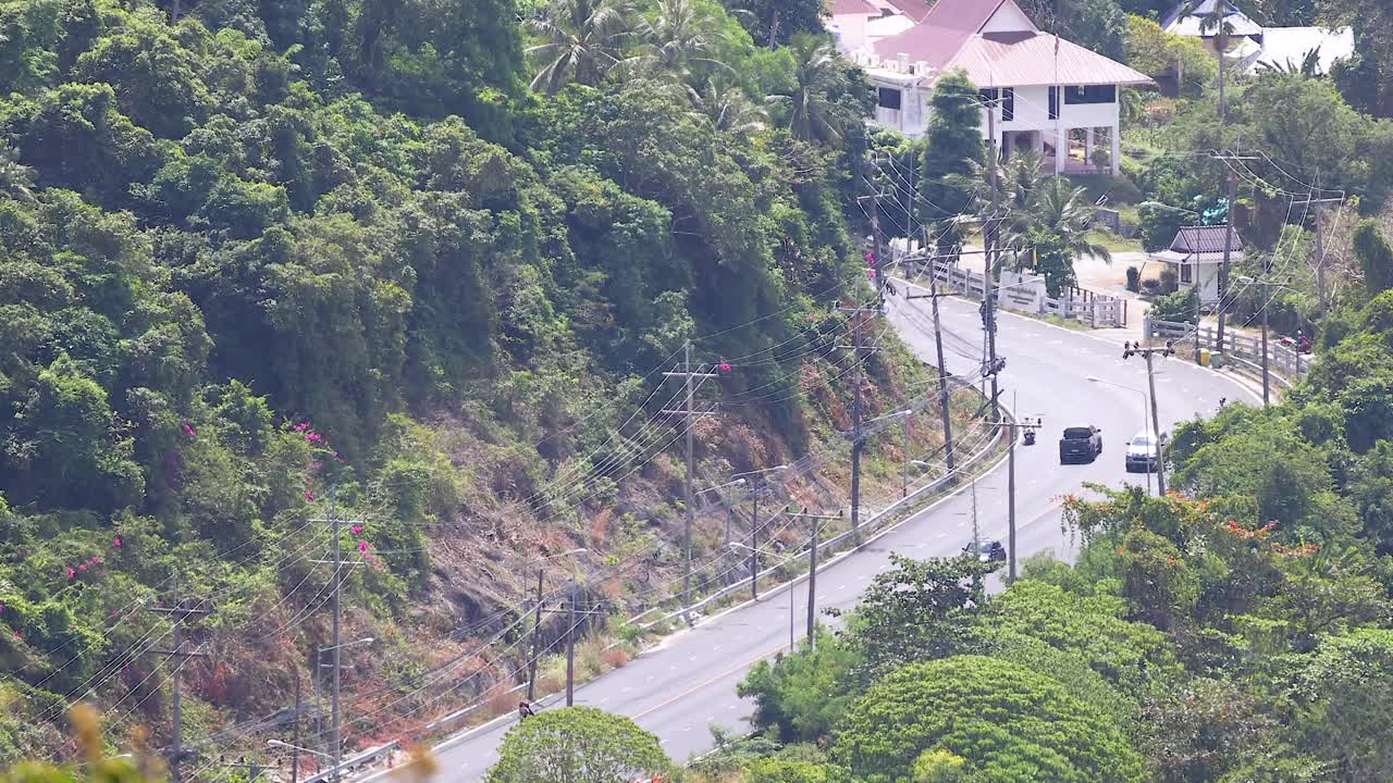 Vehicles navigate a lush, winding road in Phuket under bright daylight, highlighting the area's natural beauty and vibrant greenery
