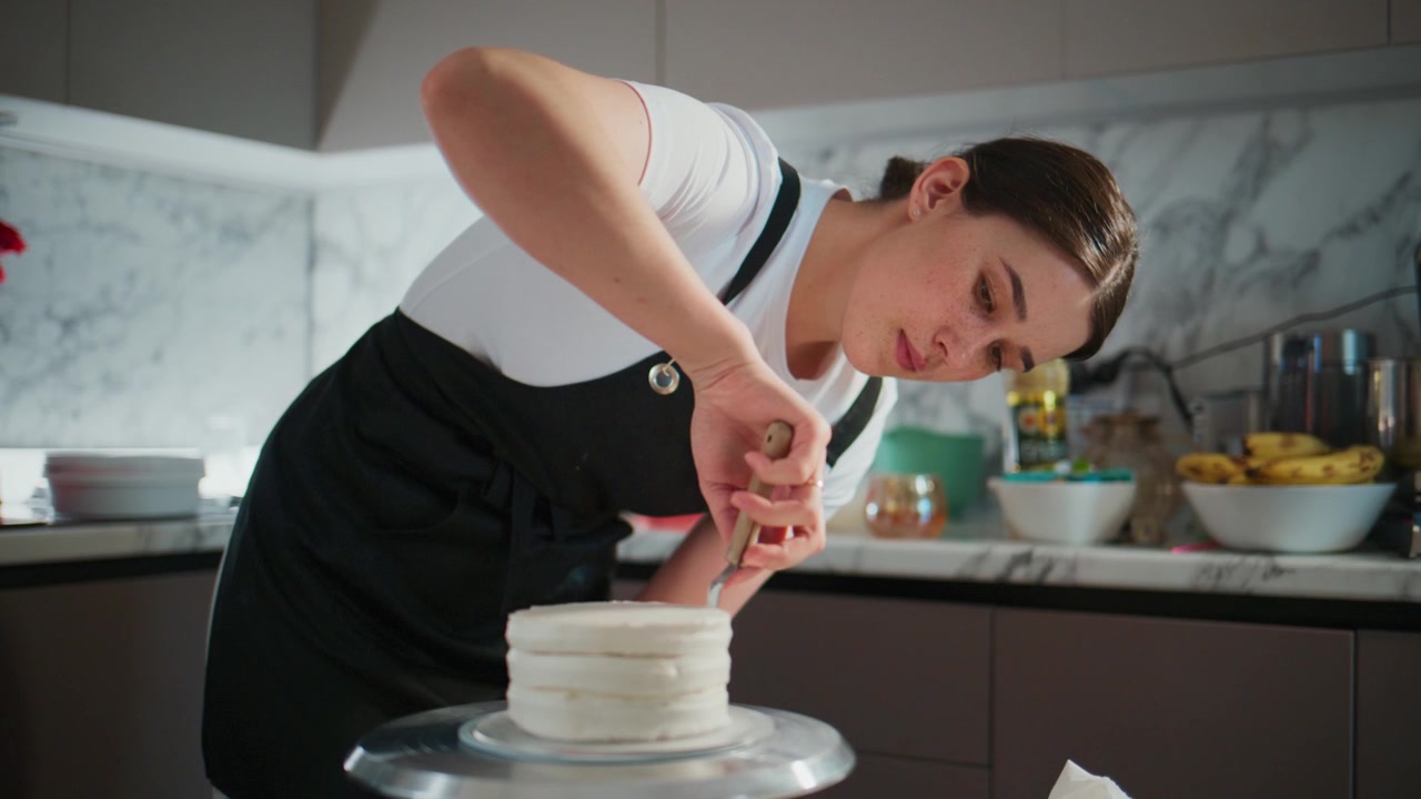 Young woman carefully decorates white cake with icing. She focuses intently on perfecting details. Kitchen has marble countertops and bright lighting. Perfect for baking, dessert
