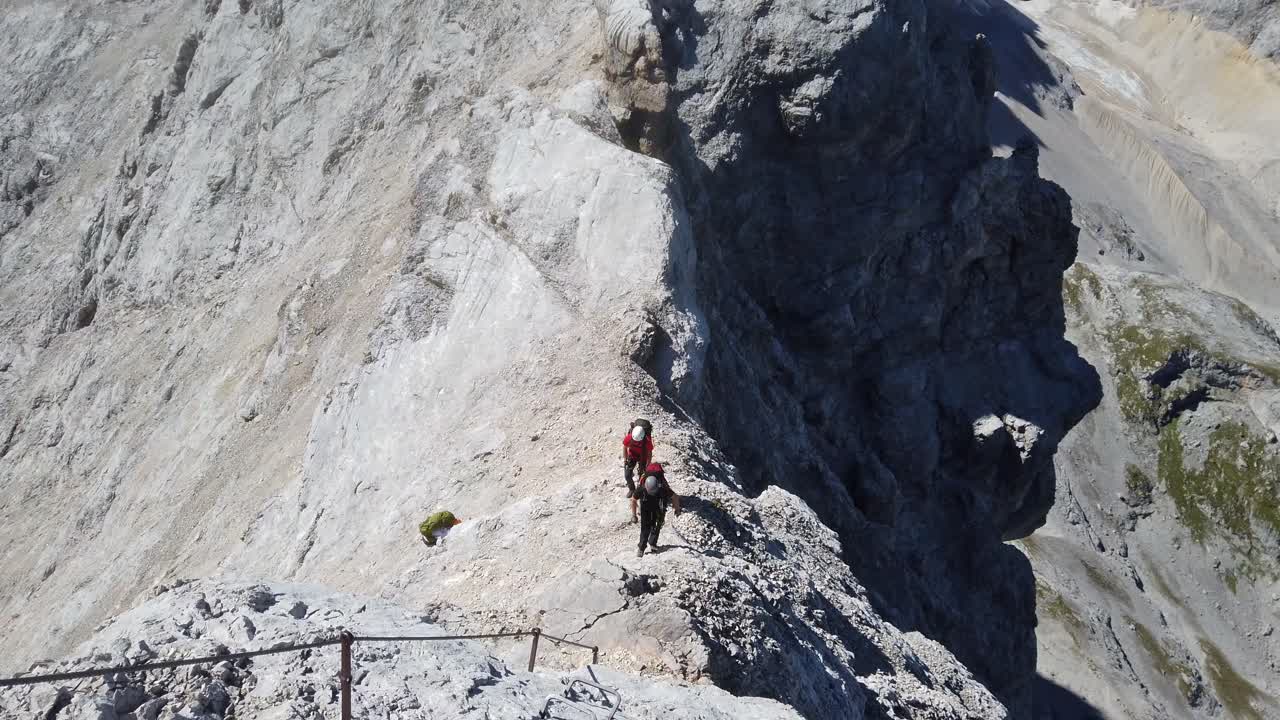 Top down pan of man climbers ascending a high alpine mountain ridge. The wear backbacks, harness and helmet and walking towards steel cables. Sunny day