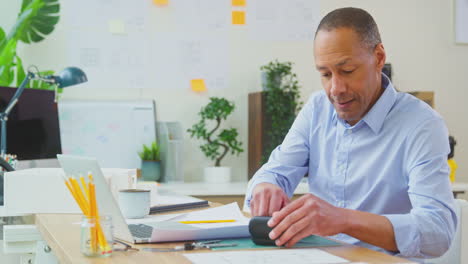 male architect in office putting in wireless earbuds working on plans for new building using laptop