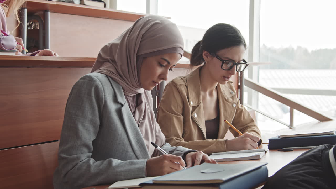Two Female Students Taking Notes in Copybooks during Lecture