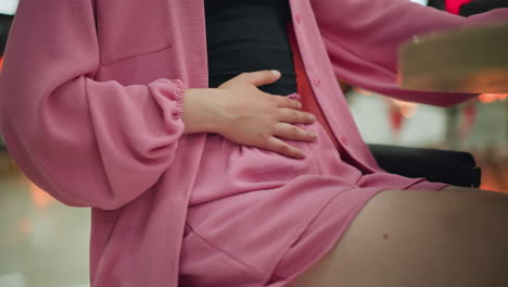 mujer con una camiseta negra bajo un vestido rosado frotando suavemente el estómago con la mano derecha, la otra mano colocada en la mesa después de una comida, el fondo presenta una luz borrosa roja