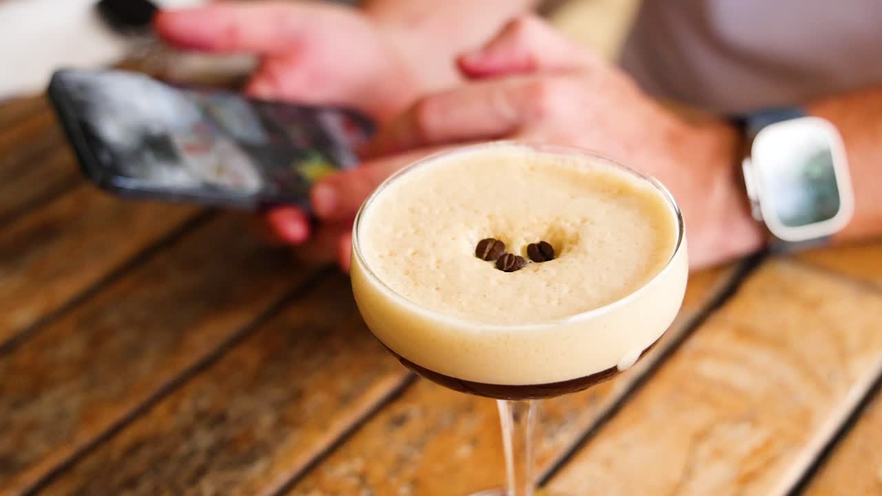 A person interacts with a smartphone beside an espresso martini on a wooden table in a well-lit cafe