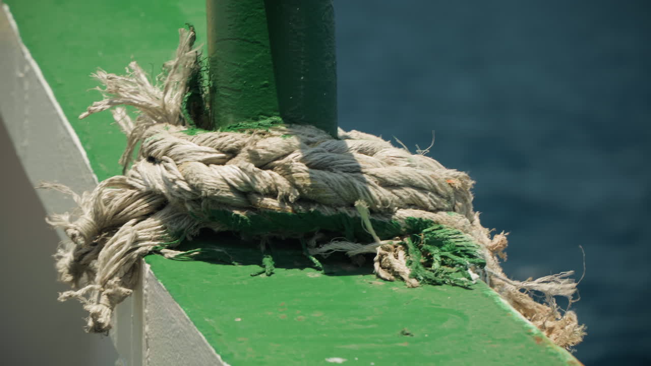 Flosset fortøjningsreb på grønt gelænder, solbelyst nærbillede af tykke vejrbidte fibre viklet omkring en stolpe med dybblå havbaggrund, teksturerede malingsflager og salt skorpe, ideel havneatmosfære.