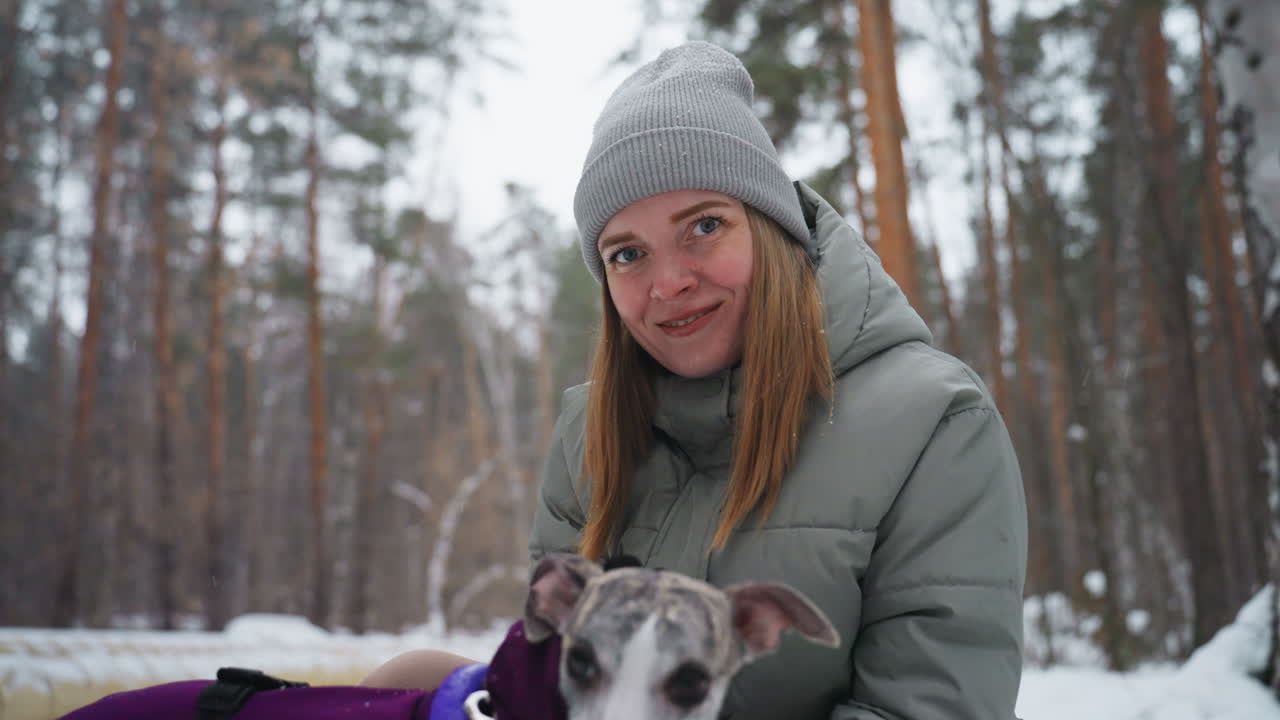Smiling woman in gray winter jacket and knit beanie sitting outdoors in snowy forest holding greyhound dog, enjoying peaceful cold weather moment, surrounded by tall pine trees in natural setting