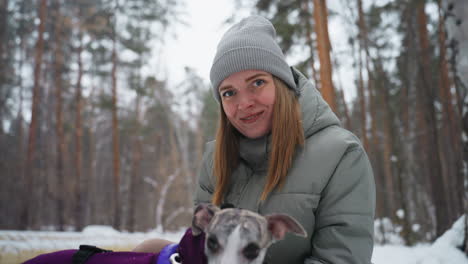 smiling woman in gray winter jacket and knit beanie sitting outdoors in snowy forest holding greyhound dog, enjoying peaceful cold weather moment, surrounded by tall pine trees in natural setting