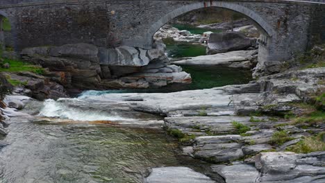 vuelo aéreo sobre el río verzasca que fluye con puente de arco en segundo plano