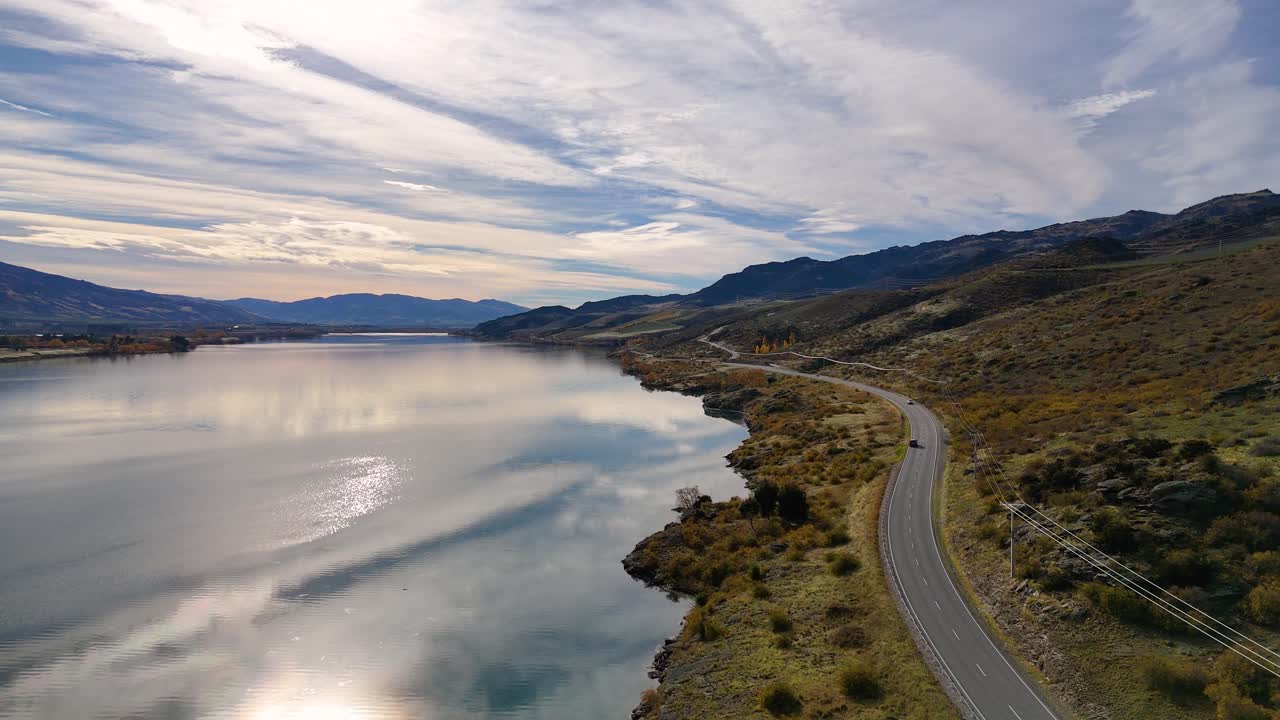 Drone footage captures serene Lake Dunstan and winding road under dramatic skies, showcasing natural beauty and tranquility