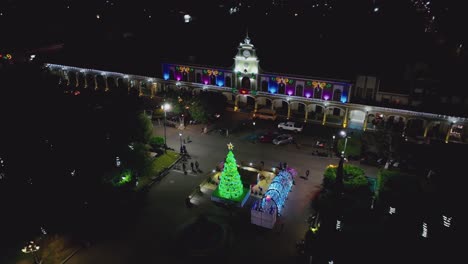 Aerial-Night-View-Of-People-Walking-At-The-Garden-Decorated-With-Glittering-Christmas-Tree-And-Lights-In-Front-Of-The-City-Hall-In-Ciudad-Guzman,-Mexico