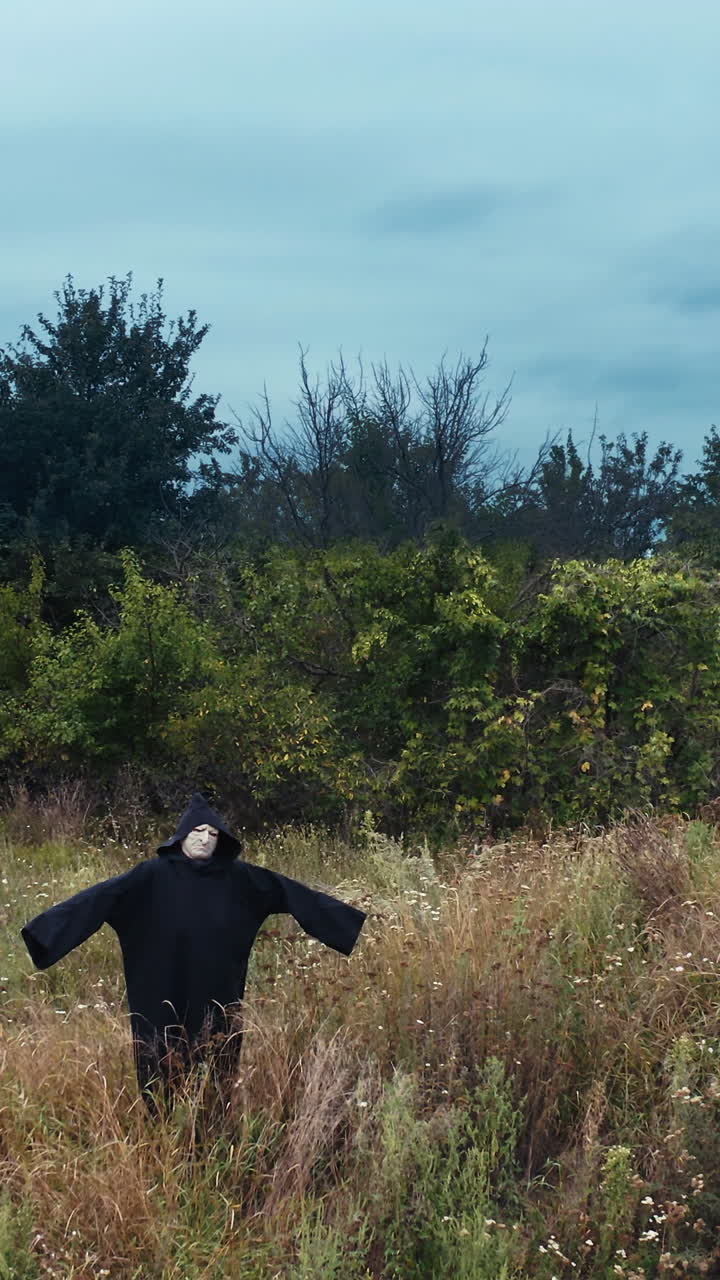 Dark ghost with outstretched arms. Scary figure of death in black mantle with hood standing on the field. Motion camera back. Halloween concept. Vertical video