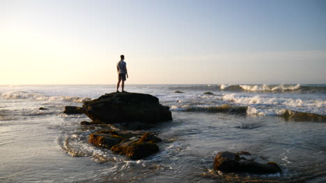 Un-Hombre-Parado-En-Una-Pose-épica-Viendo-El-Amanecer-En-Meditación-Pacífica-Mientras-Las-Olas-Del-Océano-Chocan-En-La-Playa-En-Santa-Barbara,-California-A-Cámara-Lenta