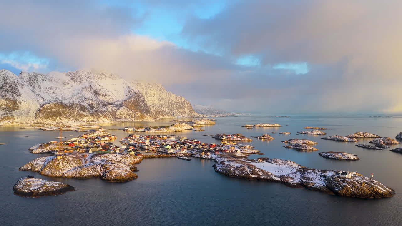 ノルウェーのロフォーテン諸島にある象徴的なヘニングスヴァー漁村のシネマティックな空撮ドローンショット。ゴールデンアワーの冬の日の出時に、雪に覆われた山々と島々が特徴的です。