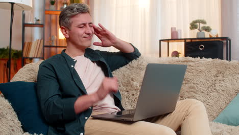 Man tired at home works on laptop massages temples with exhausted face and overwhelmed body posture