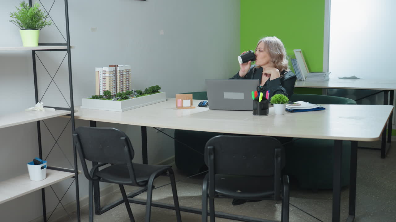 Professional lady seated in modern office setting pauses focused laptop work to sip coffee, then resumes typing beside model buildings, organized supplies, and bright green accent wall