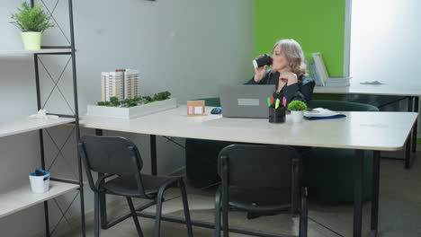 professional lady seated in modern office setting pauses focused laptop work to sip coffee, then resumes typing beside model buildings, organized supplies, and bright green accent wall