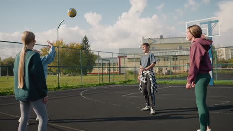 coach leads training session with three ladies passing volleyball between them on outdoor court, demonstrating teamwork and practice with basketball court and building background