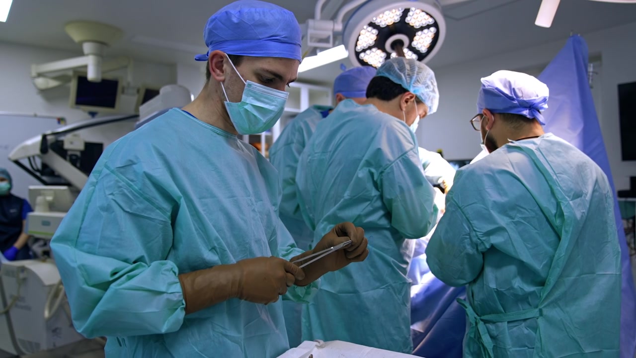 Male young Caucasian medic assisting at operation. Assistant prepares tools and materials for surgeons' usage. Doctors' team operating at backdrop.