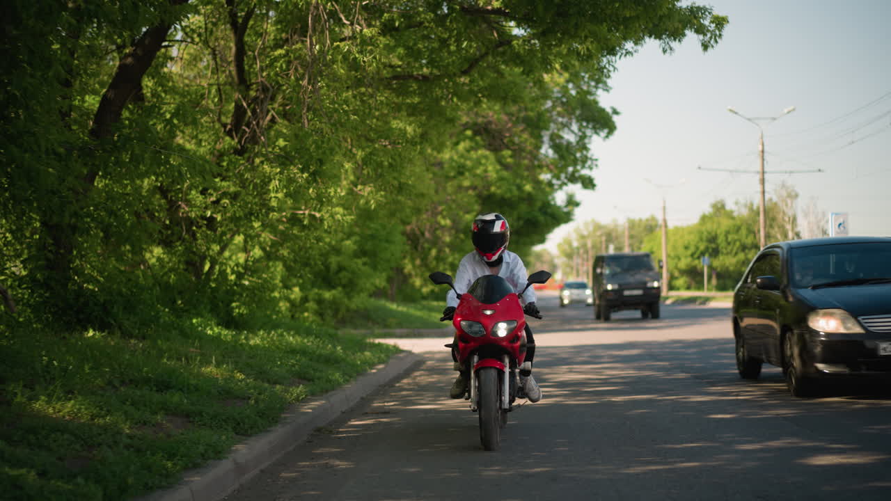 빨간색 전력 자전거를 타는 여성 오토바이 운전자는 도시 환경에서 푸른 나무 근처에 천천히 타고 있으며, 차량이 가까이 지나가며, 도로 반대편에 전기 기둥이 있습니다.