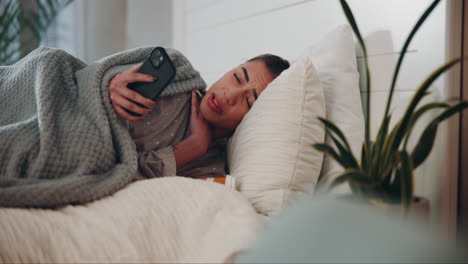 a woman lying in bed with a sore throat, holding her phone and looking worried