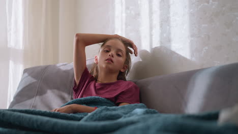 reflective girl seated on couch, hand resting on head, looking tired and unwell, showing signs of fatigue, discomfort, and illness, resting with a tired expression