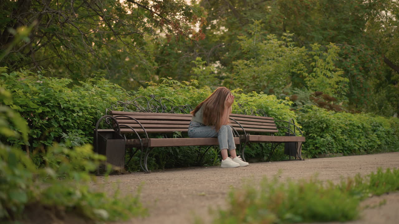 Woman seated on wooden bench in reflective, somber posture with hands gently covering her face, long blonde hair cascades down her shoulder, surrounded by lush greenery