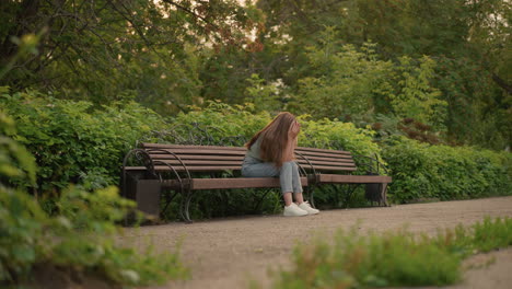 woman seated on wooden bench in reflective, somber posture with hands gently covering her face, long blonde hair cascades down her shoulder, surrounded by lush greenery