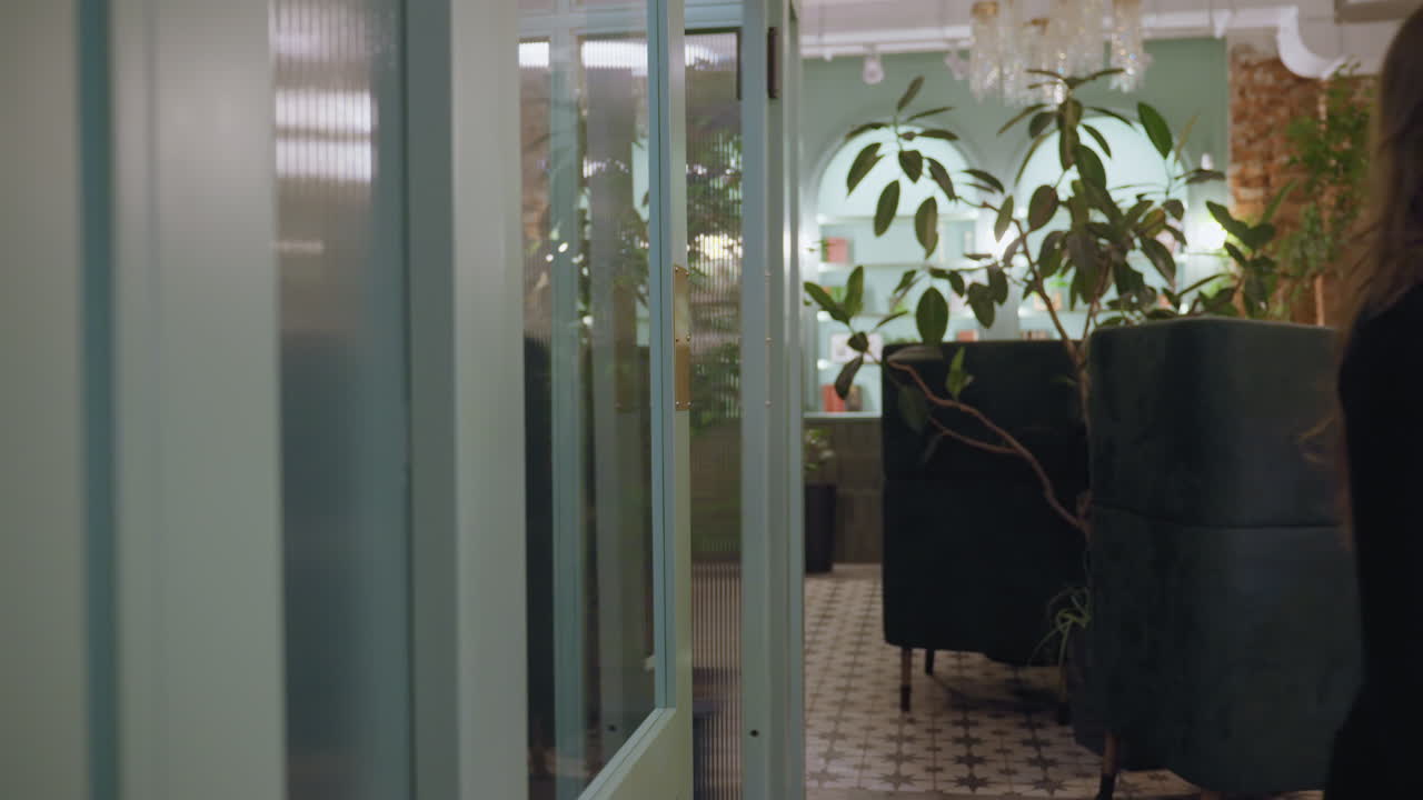 Woman pushes transparent door while entering stylish green lounge space decorated with indoor plants and cushioned seating, creating a serene and modern atmosphere in cozy interior setting