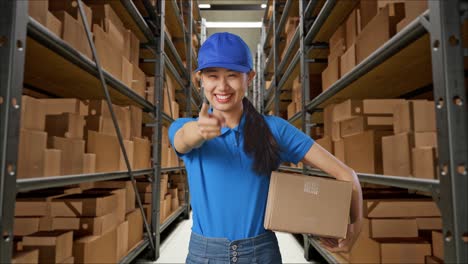 mensajera asiática en uniforme azul sonriendo y haciendo un gesto honesto mientras entrega un cartón en el almacén