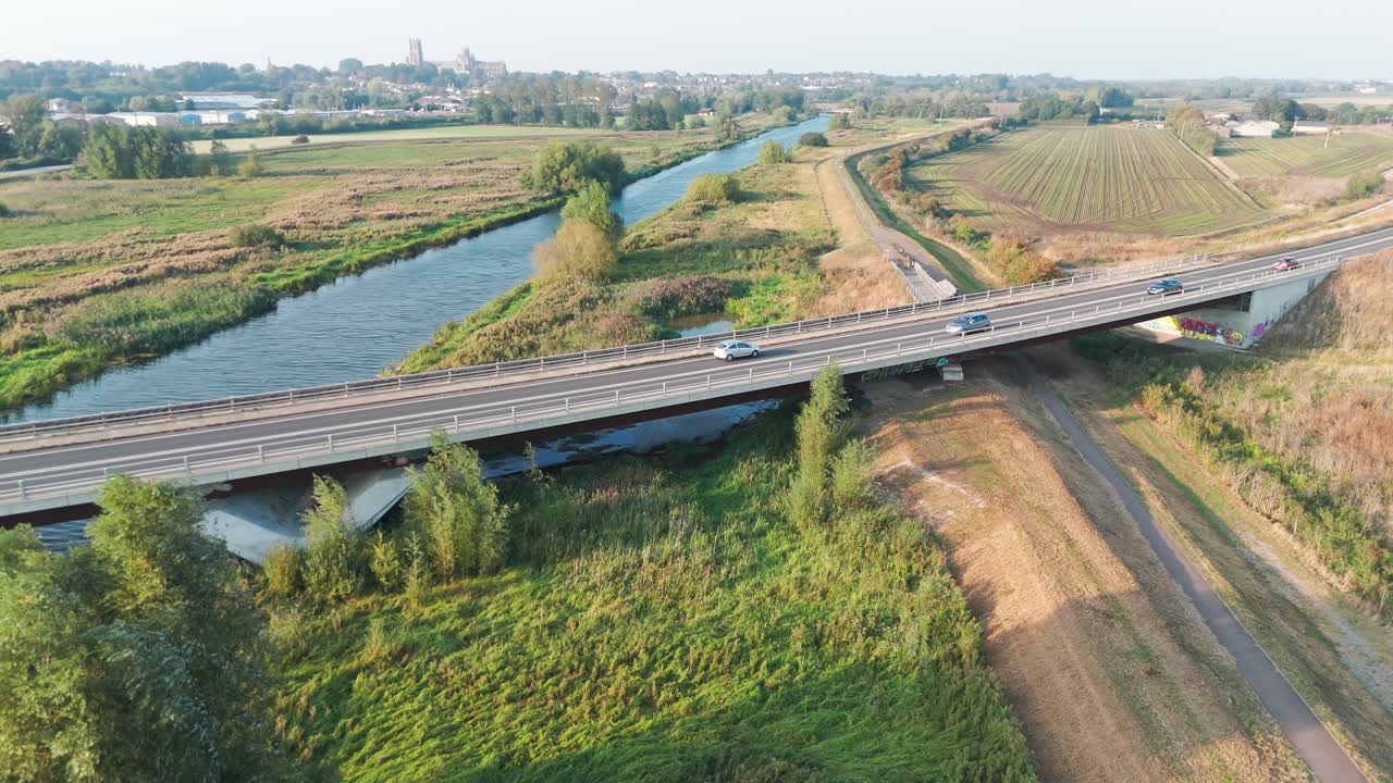 A bridge over a river with cars, surrounded by green fields and trees, under a partly cloudy sky is shot from above. The scene captures the tranquillity and beauty of rural infrastructure and nature