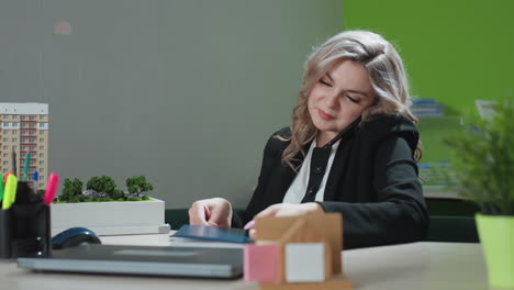 real estate agent calls client while seated in modern office, opens notebook and starts jotting notes, surrounded by model building, laptop, stationery and green potted plant on workspace