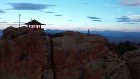 Aerial-View-of-Female-Figure-Walking-on-Top-of-Rocky-Hill-With-Fire-Spotting-Station-Building,-Colorado-USA