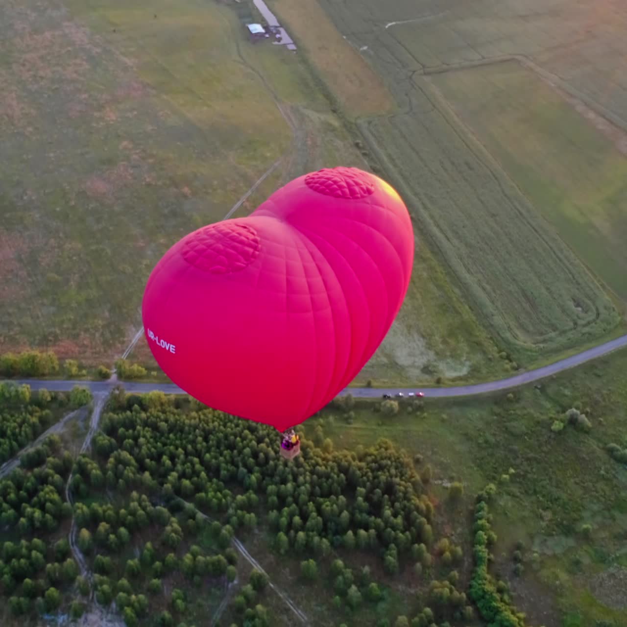 Red aerostat over fields. Romantic hot air balloon in a form of a heart travels in the sky on beautiful nature background at sunset. Top aerial view.