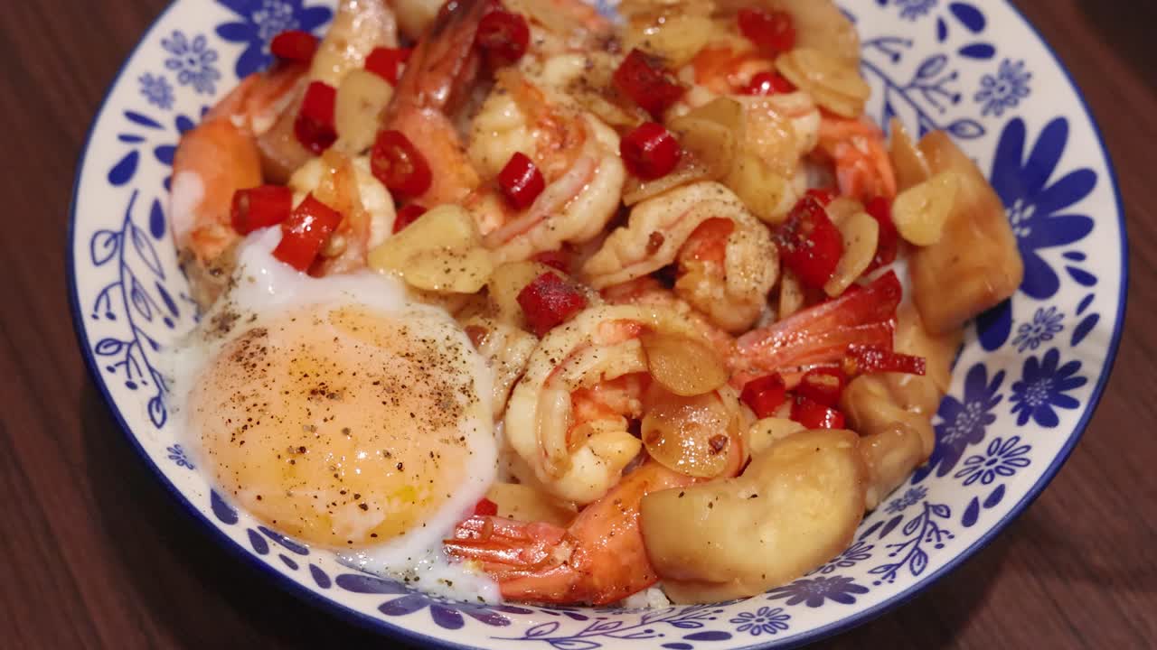 A spoon and fork break a runny egg atop Thai shrimp, garlic, chili, and rice in a patterned bowl under warm indoor lighting