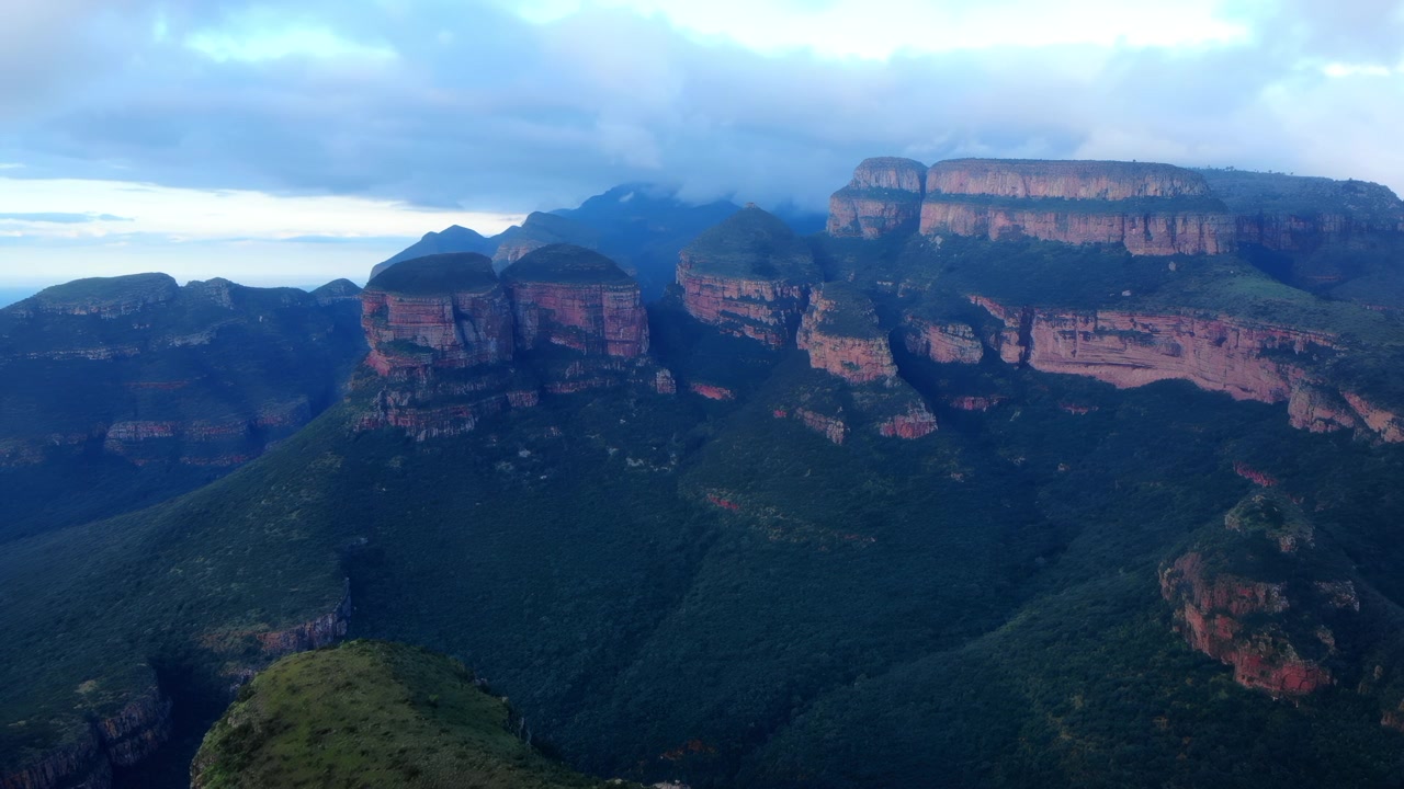 Wide panoramic view of a rugged mountain landscape with layered cliffs, a deep valley, rocky ledges, forested slopes, and a cloudy blue sky in daylight