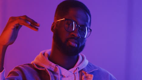 portrait of american stylish man in glasses dancing looking at camera on pink and violet neon light background