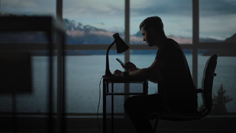 a thoughtful man sitting at a table with a desk lamp and a mobile phone. the dark day is twilight against the background of the window a sad man is thinking about work while sitting at home.