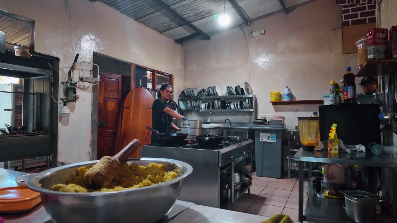 Santo Tomas, Ecuador, 1 maggio 2026: Cuoco che prepara il cibo nella cucina di un ristorante alle Galapagos. Scena interna della cucina con cuoco ai fornelli, ciotola di metallo in primo piano, scaffali, utensili e luce a soffitto.