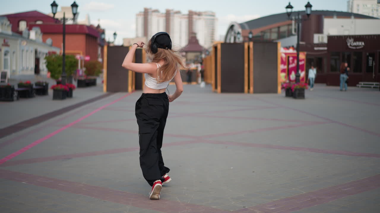 Giovane donna che balla per strada in città con cuffie e pantaloni cargo, improvvisando in una piazza vuota con giravolte giocose e passi all'indietro, scarpe da ginnastica rosse e fluenti capelli biondi ricci, casual