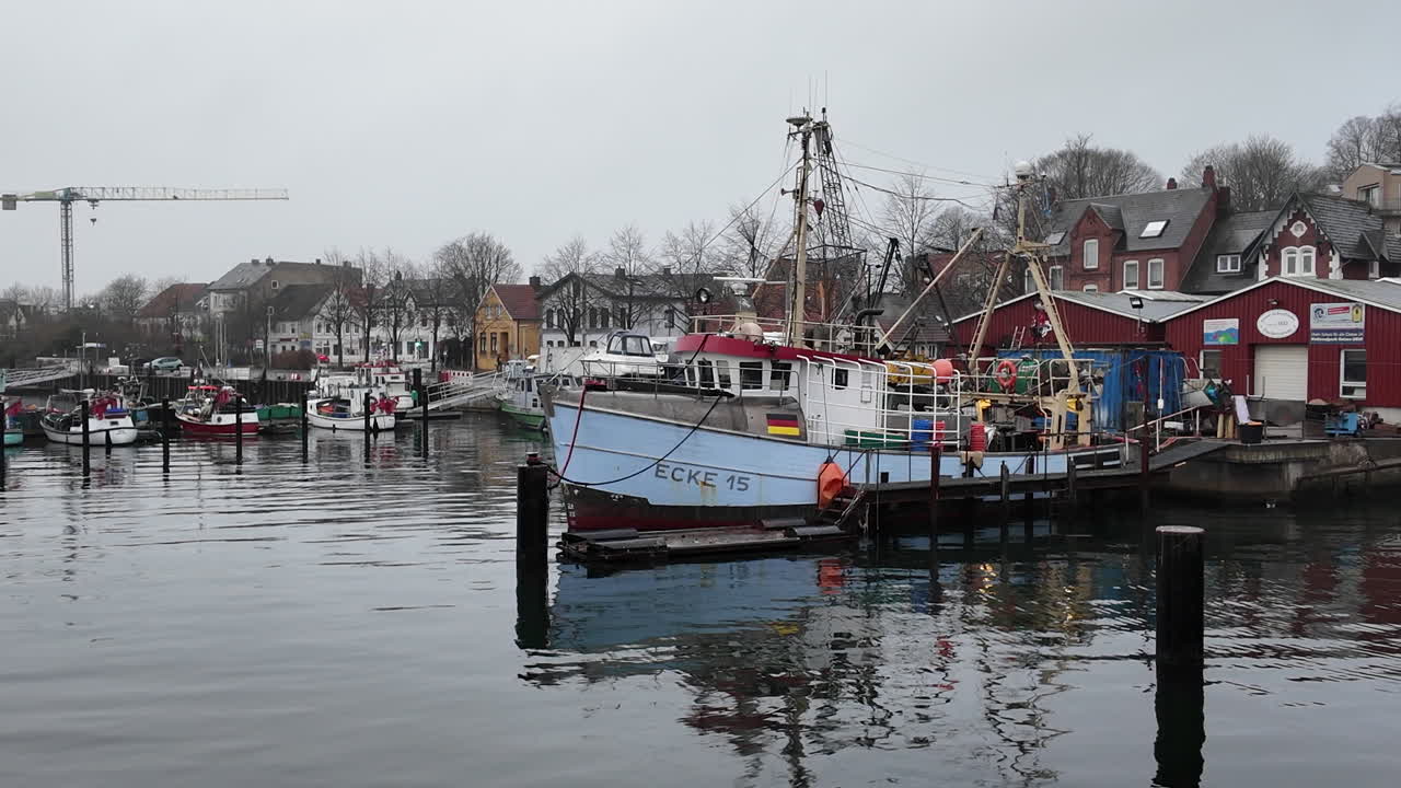 En rolig havnescene i Eckernförde, Tyskland, med tradisjonelle fiskebåter fortøyd langs vannkanten på en grå, overskyet dag. Refleksjoner skinner på det stille vannet mens historiske havnebygninger