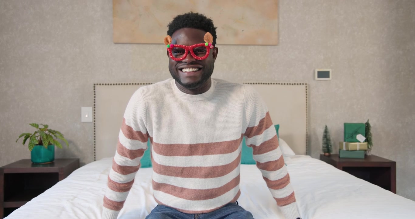 African American man laughing, lowering red glasses to offcamera cue in cream sweater jeans on bed