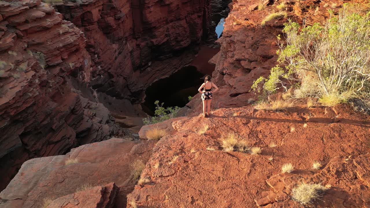 Aerial tilt down shot of pretty woman standing on edge of cliff and watching during golden sunset at Karijini National Park