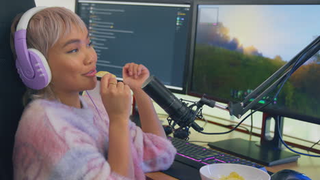 mujer comiendo bocadillos mientras juega en casa sentada en el escritorio usando auriculares para transmisión en vivo