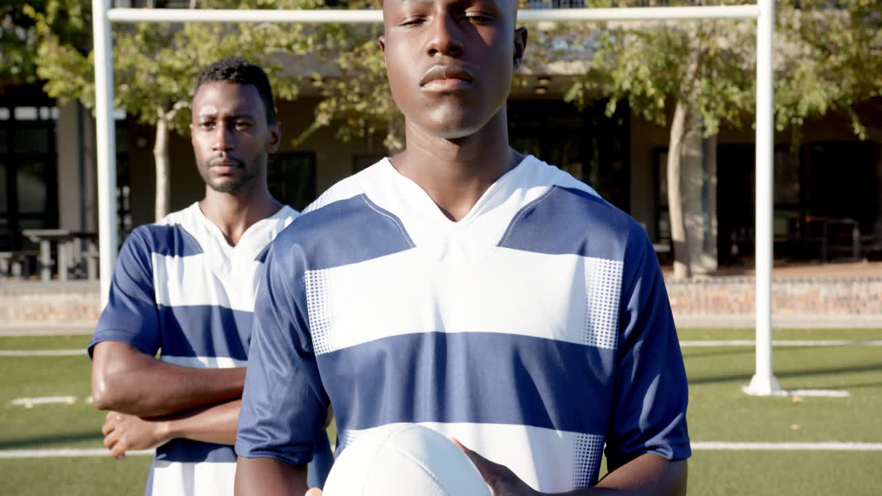 Holding rugby ball, two african american male athletes in sports uniforms standing on field
