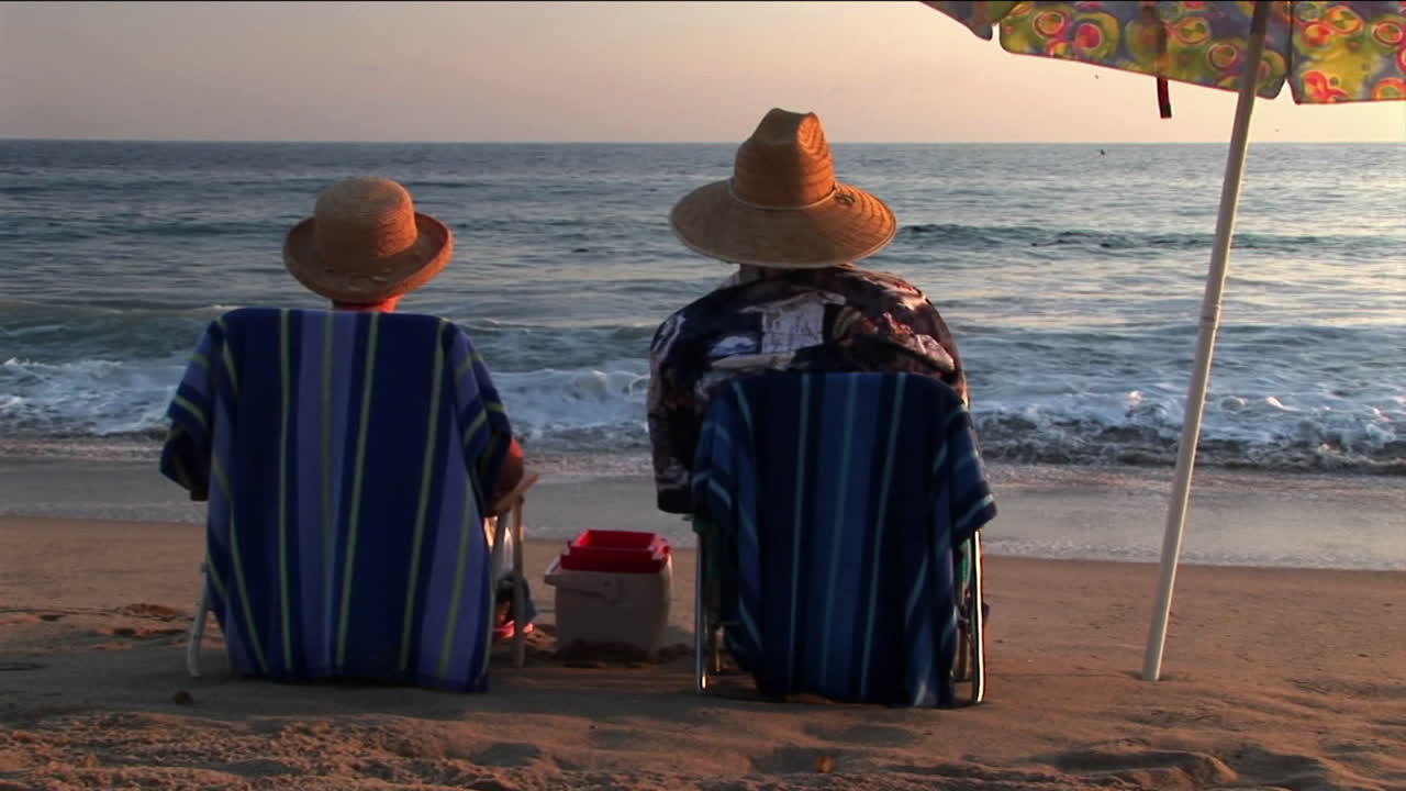 부부는 바다를 보면서 우산 아래 해변 의자에서 휴식을 취합니다.