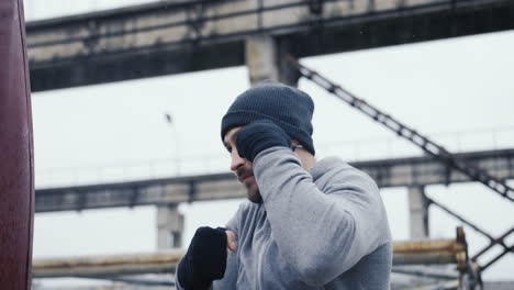 close-up view of caucasian man in grey beanie and sportswear hitting a punching bag outdoors an abandoned factory on a cloudy morning